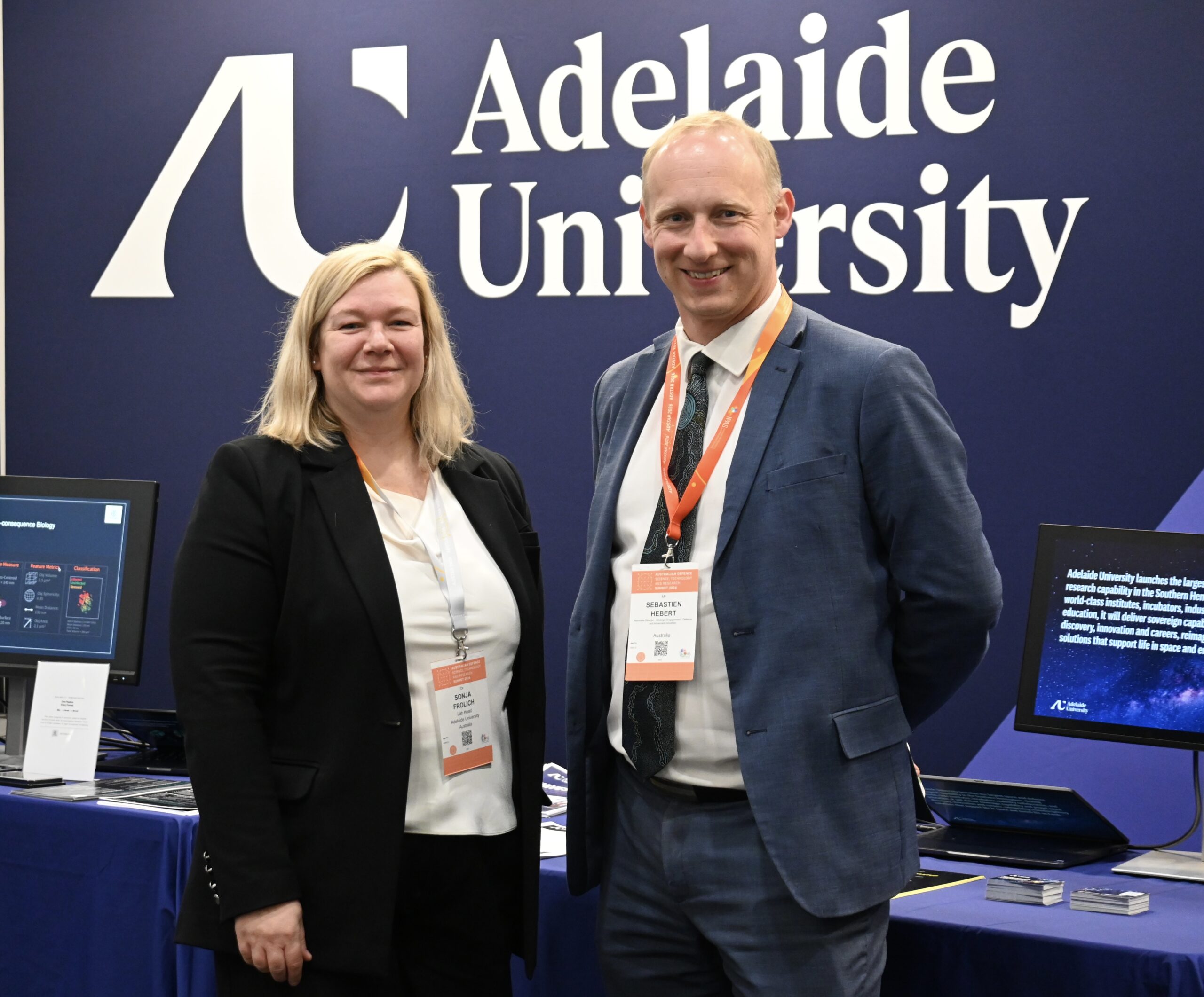 Two people looking at camera in front of Adelaide University logo